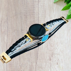 K&ouml;nnte beinhalten: Eine schwarze Smartwatch mit goldener L&uuml;nette und einem mehrstr&auml;ngigen Armband. Das Armband hat geflochtenes schwarzes Leder, ein Schlangenmuster und einen silbernen Strang, mit einem t&uuml;rkisfarbenen Steinakzent. Die Uhr liegt auf einer hellen Holzoberfl&auml;che.