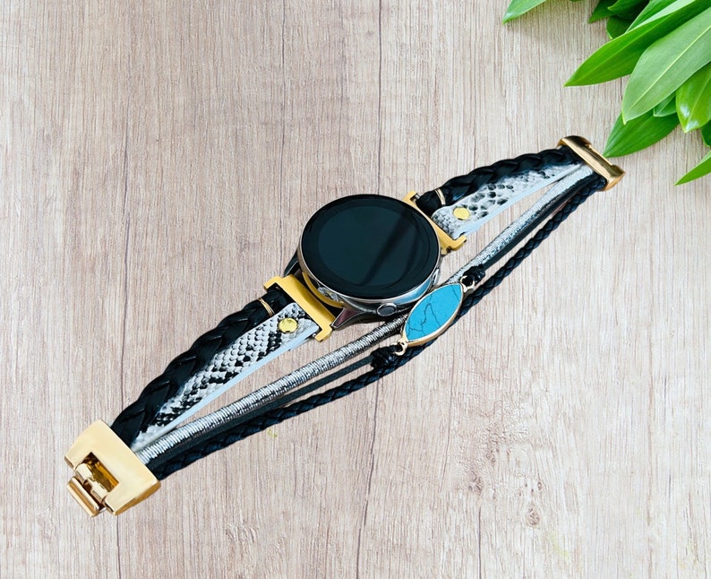 K&ouml;nnte beinhalten: Eine schwarze Smartwatch mit goldener L&uuml;nette und einem mehrstr&auml;ngigen Armband. Das Armband hat geflochtenes schwarzes Leder, ein Schlangenmuster und einen silbernen Strang, mit einem t&uuml;rkisfarbenen Steinakzent. Die Uhr liegt auf einer hellen Holzoberfl&auml;che.