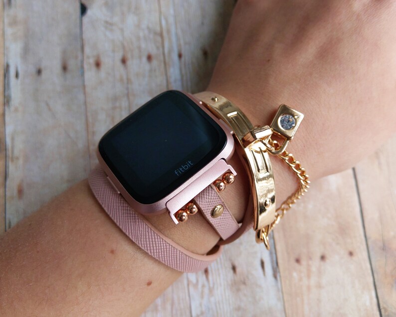 May include: A rose gold Fitbit Versa smartwatch with a black screen is worn with a matching pink leather band. A gold-colored bracelet and a gold chain bracelet with a clear gem are also worn on the wrist.