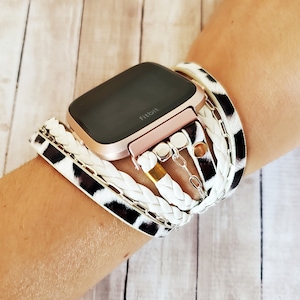 May include: A rose gold Fitbit smartwatch with a black and white animal print band and a white braided band with gold and silver charms.