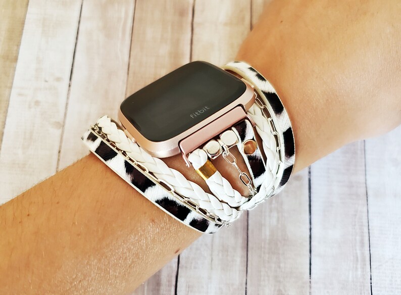 May include: A rose gold Fitbit smartwatch with a black and white animal print band and a white braided band with gold and silver charms.