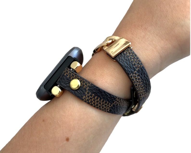 Peut inclure: Un bracelet de montre &agrave; motif noir et marron avec des accents dor&eacute;s. Le bracelet est enroul&eacute; autour d'un poignet.