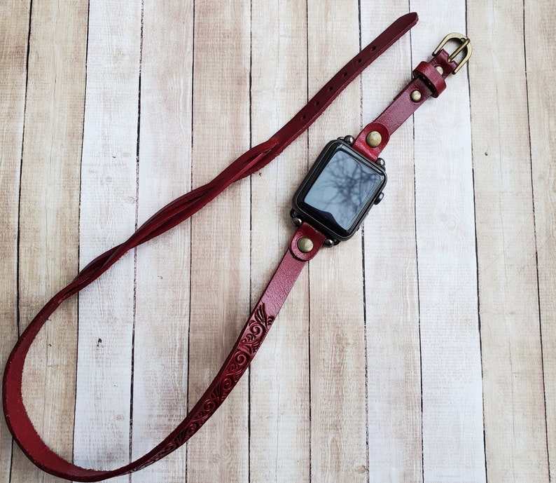 Może przedstawiać: Czerwony sk&oacute;rzany pasek do zegarka z misternym wzorem kwiatowym. Pasek jest przymocowany do czarnego Apple Watch.