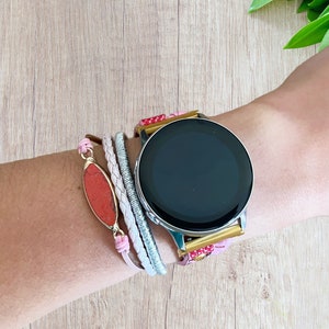 K&ouml;nnte beinhalten: Eine silberne Smartwatch mit schwarzem Bildschirm wird am Handgelenk getragen, kombiniert mit einem Armband-Stack. Die Armb&auml;nder umfassen ein rosa Lederband, ein geflochtenes wei&szlig;es Band, ein silbernes Band und einen roten Steinanh&auml;nger mit goldenem Rand.
