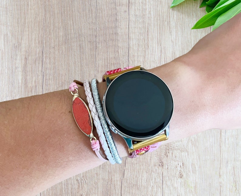 K&ouml;nnte beinhalten: Eine silberne Smartwatch mit schwarzem Bildschirm wird am Handgelenk getragen, kombiniert mit einem Armband-Stack. Die Armb&auml;nder umfassen ein rosa Lederband, ein geflochtenes wei&szlig;es Band, ein silbernes Band und einen roten Steinanh&auml;nger mit goldenem Rand.