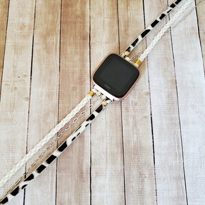 May include: A rose gold Fitbit smartwatch with a black screen is attached to a white braided strap, a black and white animal print strap, and a silver chain strap.