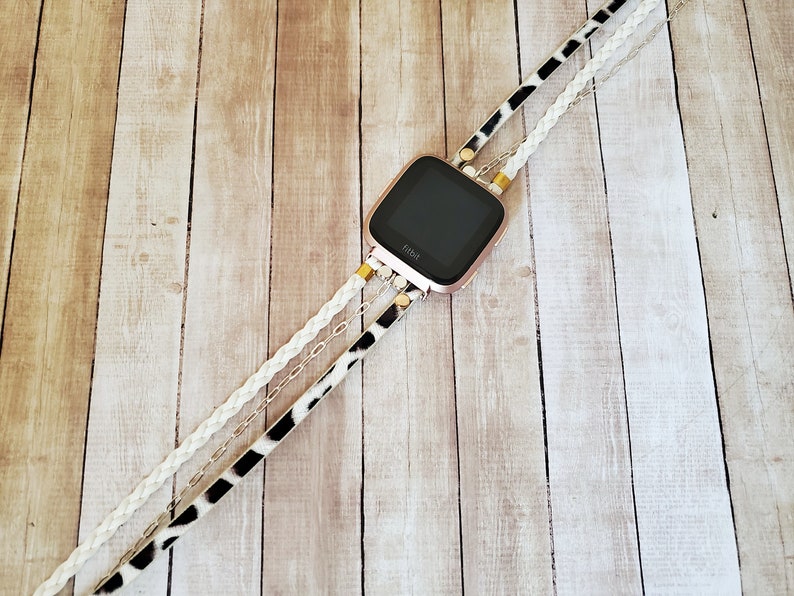 May include: A rose gold Fitbit smartwatch with a black screen is attached to a white braided strap, a black and white animal print strap, and a silver chain strap.