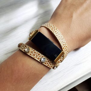 Puede incluir: Una pulsera envolvente con estampado beige y marrón con detalles dorados y adornos de cristal transparente. La pulsera se envuelve alrededor de un dispositivo rectangular negro. El diseño es una mezcla de elegancia y estilo moderno.