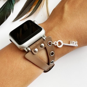 Op de afbeelding: Een bruin leren Apple Watch-bandje met een zilveren sleutelbedel met de tekst "LOVE". Het bandje heeft een driehoekig uitsparingontwerp en zilveren studs.