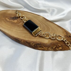 May include: A gold chain bracelet with a black rectangular charm. The bracelet is on a wooden surface.