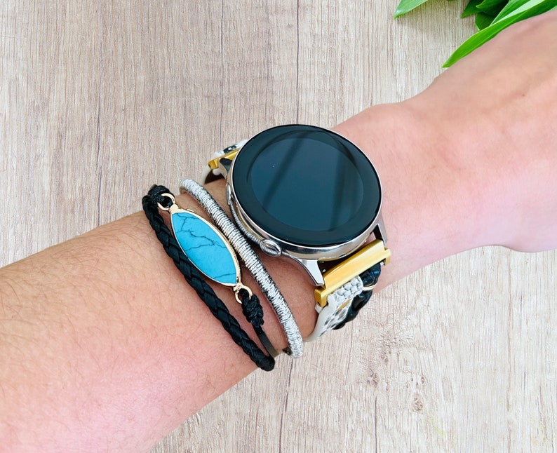 K&ouml;nnte beinhalten: Eine silberne Smartwatch mit schwarzem Bildschirm und einem Armband mit schwarzem, goldenem und wei&szlig;em Muster. Ein geflochtenes schwarzes Armband mit einem t&uuml;rkisfarbenen Stein und ein silbernes und wei&szlig;es Perlenarmband sind ebenfalls zu sehen.