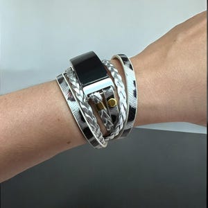 May include: A multi-layered wrap bracelet with a black rectangular watch face. The bracelet features braided and patterned straps in black, white, and silver. A gold-colored clasp secures the bracelet.