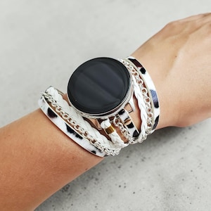 May include: A black smartwatch with a silver bezel is surrounded by a multi-strand bracelet. The bracelet features white leather, silver chain, and black and white cow print accents. The watch and bracelet are on a wrist.