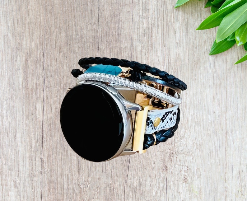 K&ouml;nnte beinhalten: Eine silberne Smartwatch mit schwarzem Bildschirm wird mit einem mehrschichtigen Armband kombiniert. Das Armband hat geflochtenes schwarzes Leder, silberne Perlen, goldene Akzente und ein Schlangenmuster.