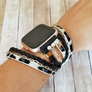 May include: A rose gold Fitbit smartwatch with a black band is worn on a wrist with a stack of bracelets. The bracelets are black leather, braided black leather, a silver chain, and a white and black animal print leather bracelet.