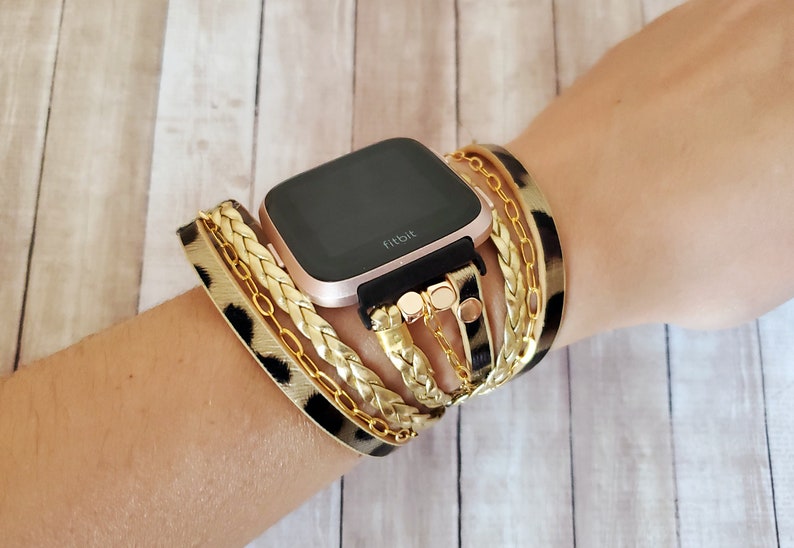 May include: A rose gold Fitbit smartwatch with a black band and a leopard print and gold chain bracelet.