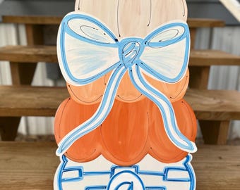 May include: A wooden pumpkin-shaped decoration with a stacked design. The top section is a light beige color, followed by orange sections, and a blue and white base. A large blue bow is painted across the center. The letter "G" is in the center of the base.