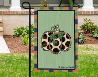 May include: A decorative garden flag featuring a pumpkin design. The pumpkin has a light green background with brown polka dots and green leaves. The flag has a green border and a multi-colored striped border. The text "THE JAGGED PAINTBRUSH" is at the bottom.