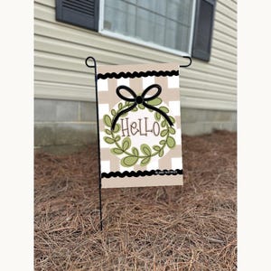 May include: A beige and white checkered garden flag with a black bow and a green wreath with the word "Hello" in the center. The flag is hanging from a black metal pole.