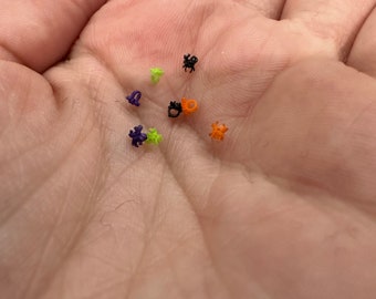 May include: A collection of seven miniature spider-shaped charms in various colors, including black, bright orange, lime green, and deep purple. Each tiny spider features a small circular loop at the top, designed for attachment to jewelry or crafts.