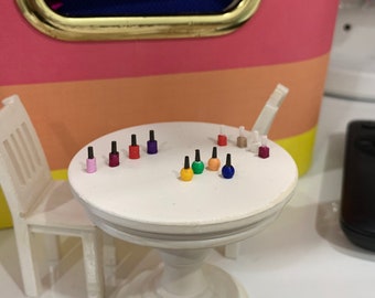 May include: A miniature white pedestal table with two matching chairs, featuring a collection of tiny, colorful nail polish bottles arranged on the tabletop. The bottles are in various shades including pink, red, purple, yellow, green, orange, blue, and white.