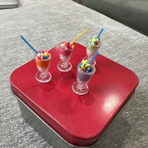 May include: Miniature dessert glasses filled with colourful layers and topped with sprinkles and straws. The desserts sit on a red, metallic surface. The glasses are clear with a stem and foot.