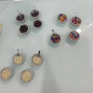 May include: A collection of miniature ceramic bowls, each filled with different colorful beads and small spoons. The bowls contain beads in various colors, including brown, rainbow, and cream, resembling cereal. The spoons are silver.