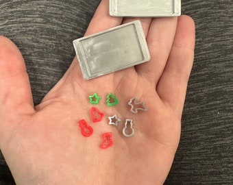 Może przedstawiać: Dwie prostokątne miniaturowe blachy do pieczenia w kolorze srebrnym oraz siedem małych foremek do ciastek o tematyce świątecznej w kolorach czerwonym, zielonym i srebrnym. Foremki mają kształty gwiazdki, choinki, dzwonka, bałwana, skarpety i renifera.