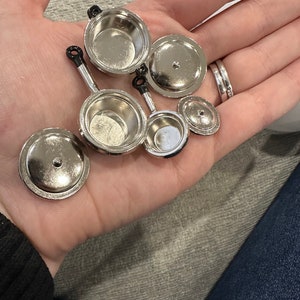 May include: Miniature silver pots and pans with black handles and matching lids. The set includes two small saucepans, a frying pan, and several lids. The shiny metal cookware is designed for a dollhouse or other small-scale project.