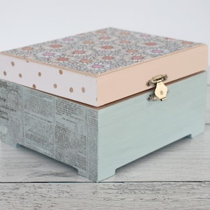 May include: A decorative wooden box with a floral daisy pattern on the lid. The lid is light pink with gold polka dots on the front edge. The box's base is light blue with a newspaper print design on the sides, and a brass clasp.