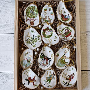 May include: A wooden box filled with twelve hand-painted oyster shell ornaments. Each shell features a unique design with gold trim, including birds, wreaths, and figures, with numbers 1 through 12. The ornaments are strung with gold cord and presented on a bed of brown paper.