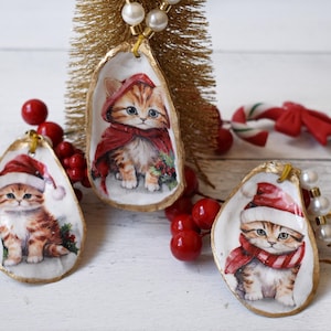 May include: Three Christmas ornaments made from oyster shells, each featuring a kitten in a Santa hat or red cape. The shells have gold edges and are strung with a pearl necklace. Red berries and a small gold tree are also in the image.