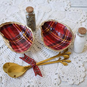 May include: Two oyster shell dishes with red plaid interiors and gold rims, alongside two small glass jars with cork stoppers, one filled with spices and the other with salt. Two gold spoons are tied with a plaid ribbon.