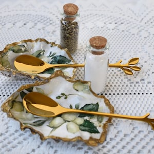 May include: Two gold spoons with leaf-shaped handles rest in white ceramic dishes with green floral designs. The dishes are on a white lace tablecloth. Two glass jars with cork tops are in the background, one filled with white powder and the other with dried herbs.