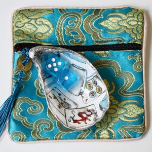 May include: A square turquoise and gold brocade pouch with a black zipper and a blue tassel featuring a round coin charm. Resting on the pouch is a white oyster shell painted with playing cards and a red dragon, containing two translucent blue dice.