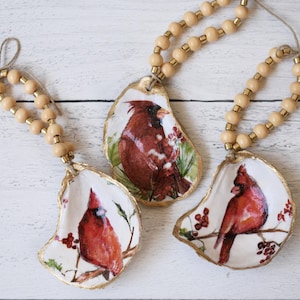 May include: Three decorative ornaments made from oyster shells, each featuring a red cardinal bird design. The shells are edged in gold and strung with wooden beads. The birds are perched on branches with berries.