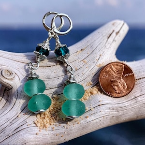 May include: A pair of handcrafted earrings featuring sea glass beads in a soft teal color. The earrings have silver-toned metal accents and lever-back closures. The earrings are displayed on a piece of driftwood with a penny for size reference.