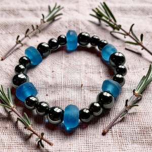 May include: A beaded bracelet featuring alternating round, polished black beads and cylindrical, matte blue beads. The bracelet is arranged in a circular pattern, showcasing a contrast between the dark, reflective spheres and the bright, opaque blue accents.