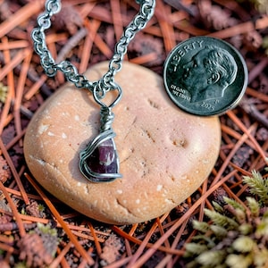May include: A wire-wrapped pendant featuring a small, deep red, raw gemstone suspended from a silver-toned metal chain. A 2025 United States dime is positioned next to the pendant to provide a clear visual reference for the scale of the jewelry.