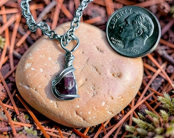 May include: A wire-wrapped pendant featuring a small, deep red, raw gemstone suspended from a silver-toned metal chain. A 2025 United States dime is positioned next to the pendant to provide a clear visual reference for the scale of the jewelry.
