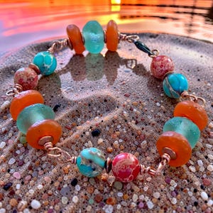 May include: A handmade bracelet featuring a variety of colorful beads. The bracelet includes orange, turquoise, and red beads, strung together with copper wire. The beads are of various shapes and sizes, creating a unique and eye-catching design. The bracelet is displayed on a sandy surface.