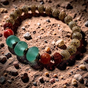 May include: A beaded bracelet featuring a variety of natural stone beads. The bracelet includes speckled, round beads, red and brown beads, and seafoam green glass beads. The beads are strung together with gold-colored metal accents. The bracelet is displayed on a sandy surface.