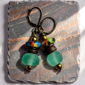 May include: Artisan earrings with a unique design. Each earring features a sea glass bead, wooden beads, and colorful glass beads, accented with gold-colored metal findings. The earrings are displayed on a gray and brown stone surface.