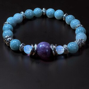 May include: A beaded bracelet featuring light blue and purple gemstones. The bracelet has a central purple bead, flanked by smaller opalescent beads and light blue beads. Silver-colored metal accents separate the beads.