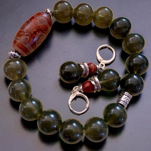May include: A beaded bracelet and matching earrings set. The bracelet features large, olive-green beads, a patterned brown bead, and silver-toned accents. The earrings have similar olive-green beads and red beads with silver-toned hooks. Jewelry.