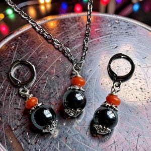 May include: A jewelry set consisting of a necklace and two matching earrings. Each piece features a large, round, dark gray metallic bead topped with a smaller, vibrant orange bead and silver-toned decorative metal caps on a silver chain.