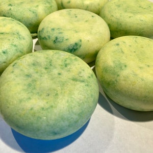 May include: Close-up of six round, pale green and yellow soap bars with a marbled appearance. The soap bars are arranged in a semi-circle on a white surface.