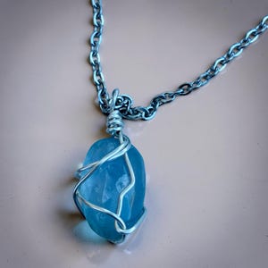 May include: A handmade necklace featuring a light blue, irregularly shaped stone pendant wrapped in silver-colored wire. The pendant hangs from a silver-colored chain, creating a simple yet elegant design. The chain is approximately 20 inches long.