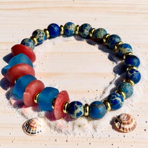 May include: A beaded bracelet featuring a mix of round blue and green marbled stone beads, translucent red and blue sea glass discs, and small gold-toned metal spacers arranged in a circular pattern on a light-colored wooden surface.