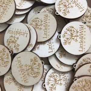 May include: A pile of round, wooden tags with the text "Little & Wild - Flowers" printed on them. Each tag features a small, stylized illustration of a flower.